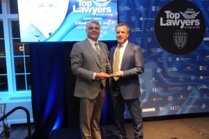 Two men in suits on a stage, holding a glass award, with a blue backdrop reading 'Top Lawyers of Long Island' in the background.