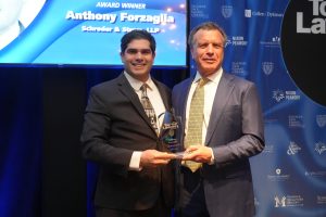 Two suited men on stage shake hands and pose with a glass award in front of a blue backdrop and screen showing the name Anthony Forzaglia (award winner).