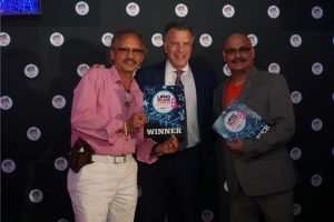 Three men posing at an awards ceremony, with the center man in a suit holding a 'WINNER' plaque while the others hold certificates.