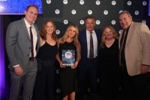 Six adults in formal attire pose together at an awards event; the center woman holds a WINNER plaque with a blue starry design and backdrop.