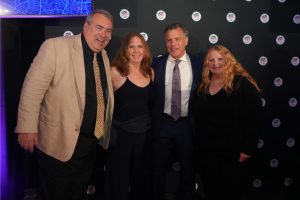 Four adults in formal attire posing and smiling for a photo against a backdrop covered in circular event logos.