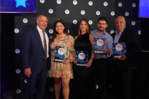 Five people pose on a stage holding 'WINNER' certificates at an award ceremony, with a branded backdrop behind them.