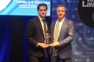 Two men in suits pose on stage holding a glass award together, with a blue backdrop behind them.