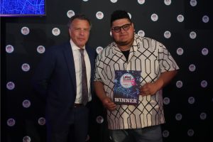 Two men pose on a black backdrop at an awards event; the man on the right holds a 'WINNER' certificate.