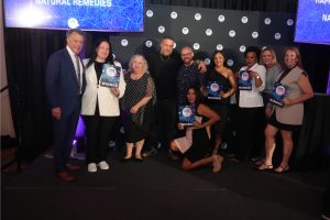 Group of diverse winners on a stage posing for a photo, holding award certificates that read WINNER, with a blue backdrop that says NATURAL REMEDIES.