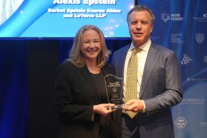 Two adults in formal attire stand on stage, smiling while presenting a glass award to each other at a professional event with a blue backdrop.
