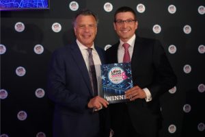 Two men in suits posing together at an awards event, holding a glossy WINNER certificate amid a backdrop of circular logos.