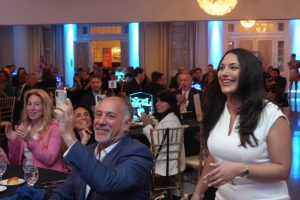 Man in a blue suit takes a selfie with a smiling woman in a white dress at a formal gala, chandeliers glowing overhead.
