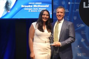 Man in a suit and a woman in a white dress pose on stage, holding a glass award; backdrop and screen read 'Award Winner Brooke McDonald'.