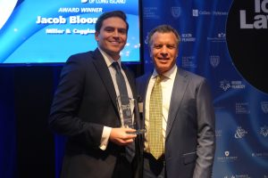 Two men in suits posing with a glass award at a formal awards ceremony, against a blue sponsor backdrop.