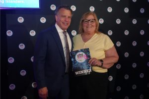 Man in a blue suit and tie stands with a smiling woman in a pale yellow top, holding a 'WINNER' award plaque at a Long Island event backdrop.