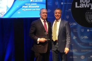 Two men in suits posing on stage at a law awards event; one holds a glass trophy in front of a blue sponsor backdrop.