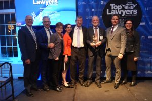 Group of eight professionals in suits posing on stage at an awards event, holding a glass trophy, with 'Top Lawyers of Long Island' backdrop.