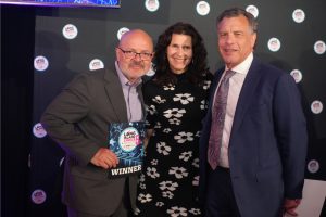 Three people pose on a stage at an awards event; the man on the left holds a WINNER plaque, flanked by a woman and a man in a suit.