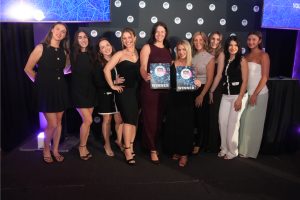 Line of ten women in evening attire pose on a stage, two centered winners hold certificates that read WINNER.