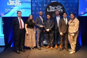 Seven professionals in formal attire pose on a stage at a Top Lawyers awards event, one man holding a glass trophy in front of a blue backdrop with logos.