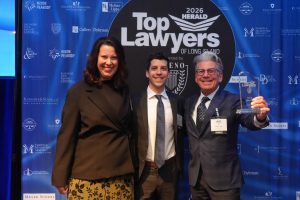 Three smiling professionals pose for a photo at a legal awards event in front of a blue backdrop reading 'Top Lawyers of Long Island'; the man on the right holds a glass trophy.