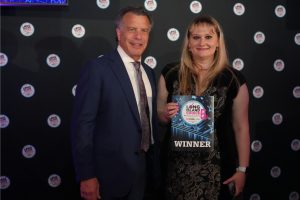 Man in a navy suit with patterned tie and a woman in a black dress hold a WINNER award plaque at an awards event backdrop.