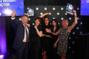 Group of five adults on stage celebrating, two women hold up 'WINNER' signs amid a branded backdrop, thumbs up and smiles all around.