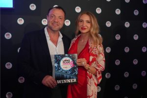 Pair poses with a winner's plaque at an awards event; man in a dark blazer and woman in a red dress hold a sign that says WINNER.