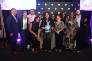 Group of 12 people posing on stage, center person holding a 'WINNER' certificate at an awards event with branded backdrop and purple lighting.
