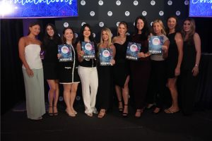 Group of women on stage holding 'WINNER' certificates at an awards event, smiling for the camera.