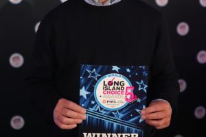 Smiling man in a dark sweater holds a blue 'WINNER' award plaque at the Long Island Choice Awards, with a starry backdrop.