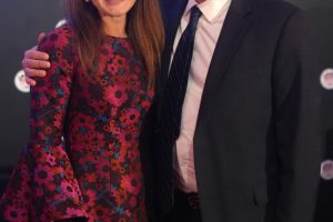 Smiling man in a suit and tie with a woman in a red floral dress posing arm in arm at a formal event backdrop with logos.
