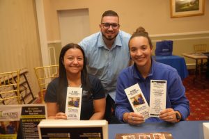 Three smiling adults seated at a conference booth, holding therapy flyers and brochures on a table.