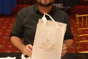 Smiling man with dark hair and glasses sits at a table, holding a white Amber Court gift bag at an event.