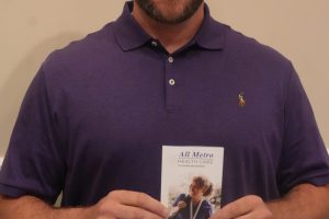 Smiling man in a purple polo holds a health-care brochure against a beige wall.