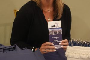 Smiling blonde woman in a black top seated at a table, holding a purple brochure with the Jerusalem Memorial Chapels logo.