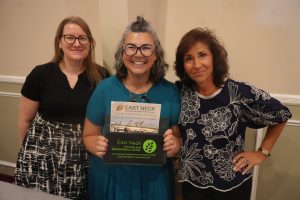 Three women smiling at the camera; center woman holds an East Neck Nursing and Rehabilitation Center brochure.