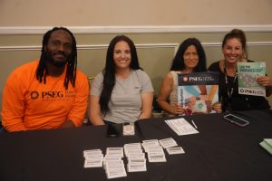 Four people sit at a table with PSEG Long Island materials, smiling at the camera; several booklets are visible, including a Sudoku book held by the person on the right.