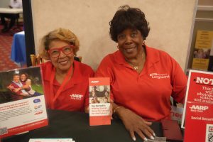 Two smiling women in red AARP Long Island shirts sit at a table with brochures and posters at an information booth.