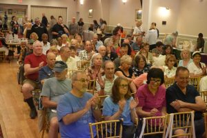 Crowd of mostly older adults seated on gold chairs in a community hall, clapping during an event.