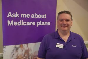 Man in a purple polo smiling beside a purple banner that reads 'Ask me about Medicare plans'.