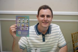 Man in a striped polo holds up a colorful brochure displaying health products. The person sits at a table, showing a product flyer to the camera.