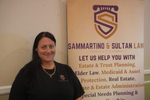 Smiling woman in a black shirt stands beside a tall banner for Sammartino & Sultan Law with a purple and orange logo.