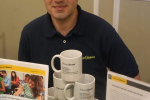 Man seated at a display table with Edward Jones brochures and mugs, wearing a dark polo shirt and looking at the camera.