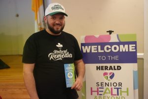 Bearded man in a black shirt and white cap with 'Natural Remedies' logo standing beside a colorful 'WELCOME TO HEALTH & BEYOND' sign poster.