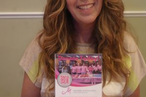 Smiling woman with long wavy hair holding a pink and white fundraising flyer at a table.