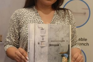 Woman smiling and standing, holding a Bath Solutions brochure with bathroom fixtures illustrated on the cover in front of a display wall.