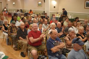 Audience of seniors seated on gold chairs in a banquet hall, clapping during a community event.