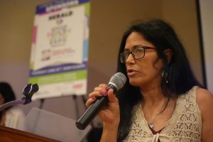 Woman with glasses speaks into a handheld microphone at a podium, poster in a blurred background.