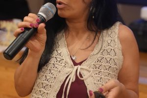 Woman speaking into a handheld microphone, wearing glasses and a cream crocheted vest over a burgundy dress at an event