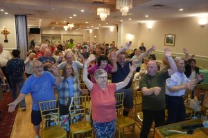 A large group of people in a banquet hall with their arms raised in celebration or cheering at a community event.