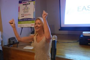 Smiling woman on stage raises her arms in celebration, with an expo poster behind her and a microphone in the foreground.