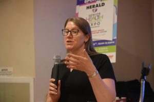 Woman with glasses speaking into a handheld microphone at an indoor event, gesturing with her hand in front of a banner backdrop.