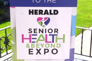 Outdoor signboard on a metal easel saying 'WELCOME TO THE HERALD SENIOR HEALTH & BEYOND EXPO' with colorful graphics near a brick wall and green lawn in the background